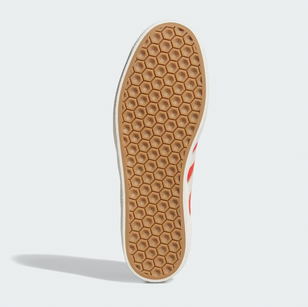 The image showcases the sole of an ADIDAS GAZELLE ADV BETTER SCARLET / CHALK WHITE shoe, displaying a honeycomb pattern in a light brown hue against a pristine white backdrop.