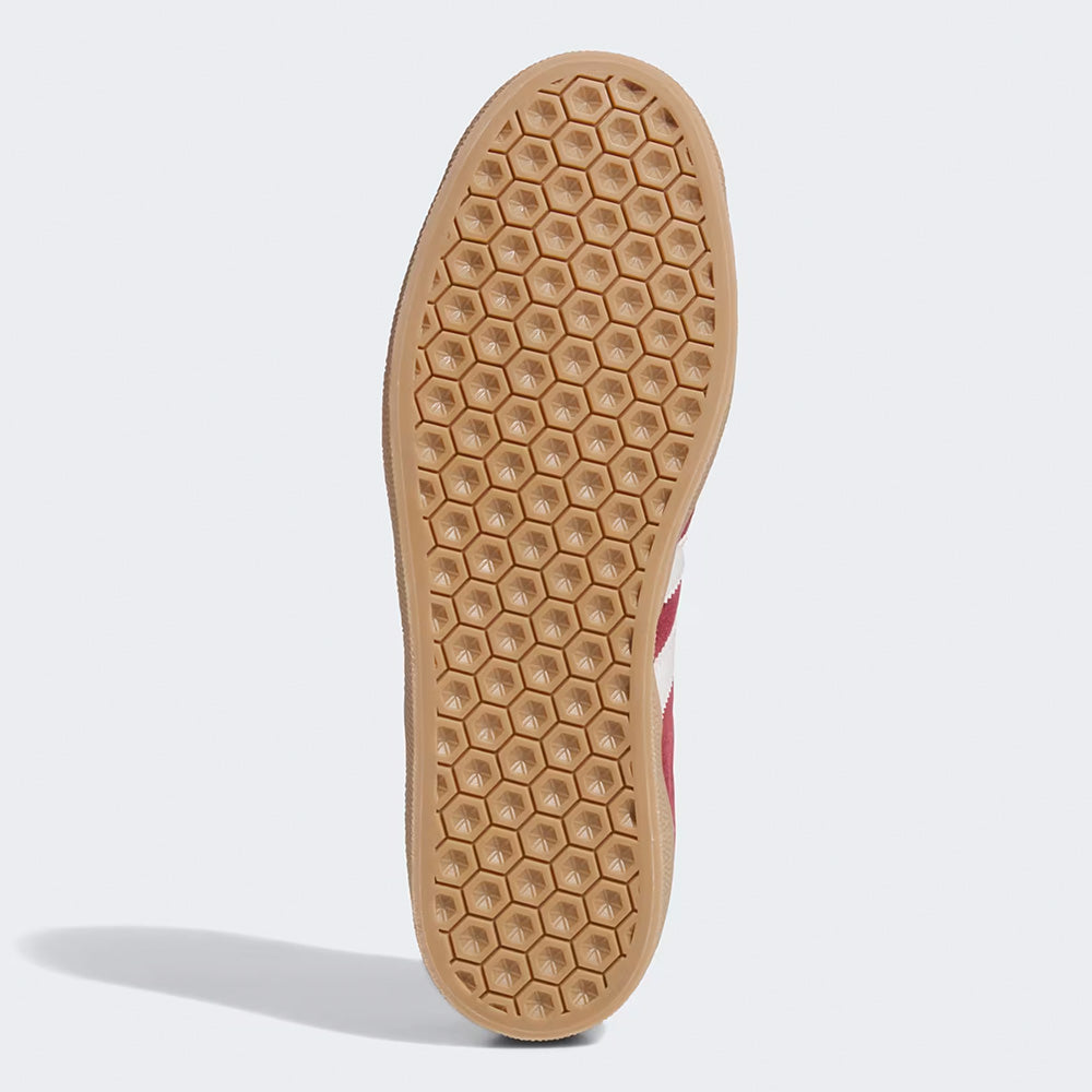 The image shows the sole of the ADIDAS GAZELLE ADV in Shadow Red, Crystal White, and Gold Metallic, featuring a honeycomb-patterned tread and light brown color, highlighting its durability as a skate shoe.