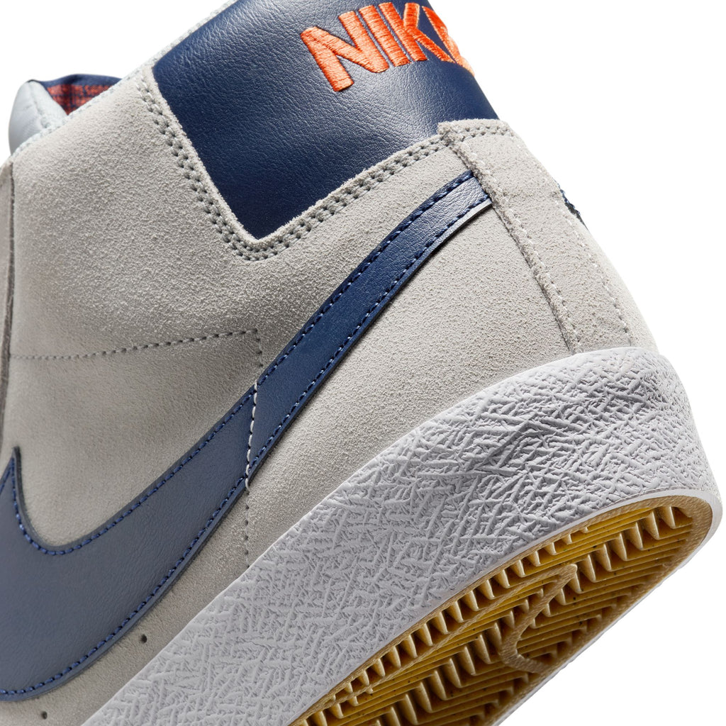 Close-up of the heel of a NIKE SB BLAZER MID WOLF GREY / MIDNIGHT NAVY sneaker, featuring an orange logo, textured sole detail, intricate stitching, and Zoom Air cushioning. Ideal for skateboarders who prioritize both style and performance.