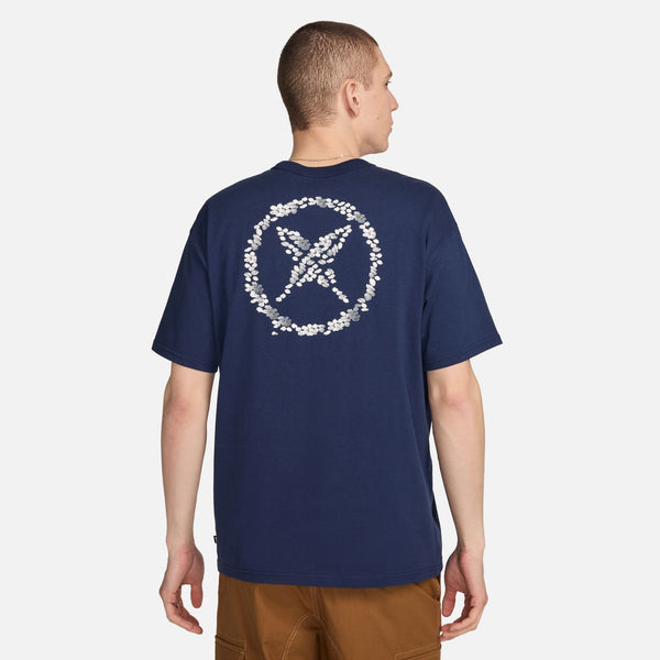 Man in a navy blue Nike Men's Skate T-Shirt with a white floral peace sign design on the back, standing turned slightly to the side.