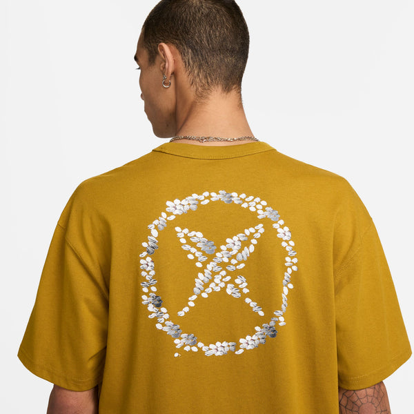 Rear view of a person wearing a NIKE SB YUTO TEE BRONZINE men's skate t-shirt with a white floral peace symbol design on the back.