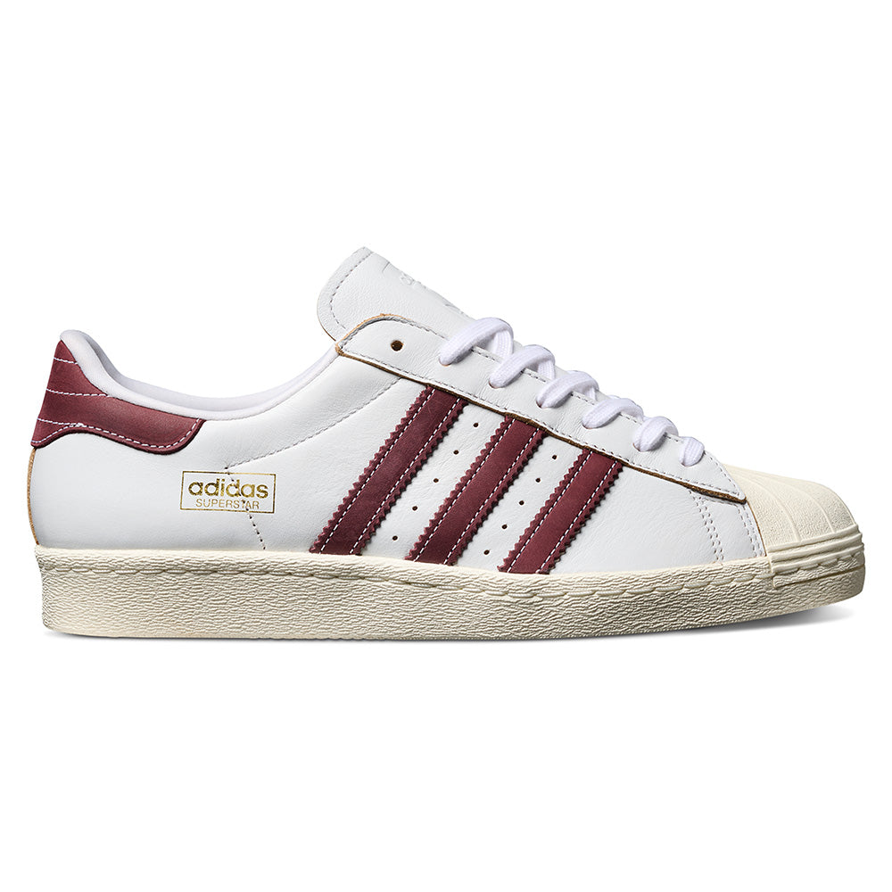 The ADIDAS SUPERSTAR 80 ADV in Crystal White/Shadow Red features maroon stripes and heel tab, suede upper, off-white sole, flexible cupsole, and gold Adidas branding on the side.