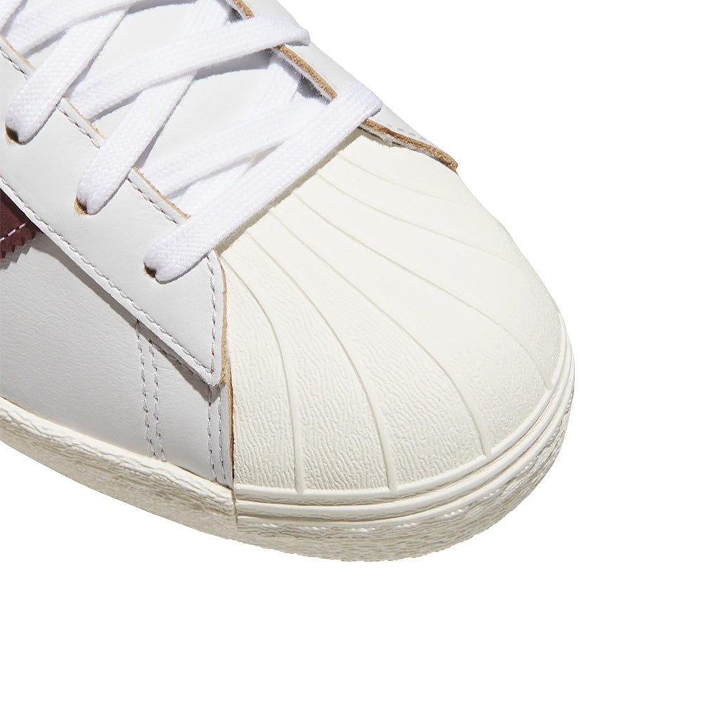 Close-up of the ADIDAS SUPERSTAR 80 ADV sneaker in Crystal White/Shadow Red, featuring white laces and a textured rubber toe cap.