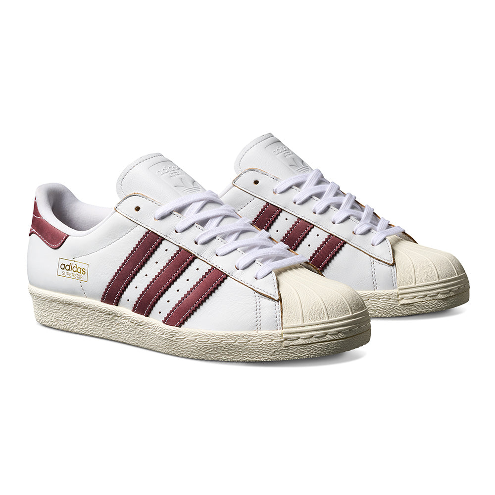 The ADIDAS SUPERSTAR 80 ADV in Crystal White/Shadow Red features maroon stripes, beige rubber toe caps, and textured off-white soles.