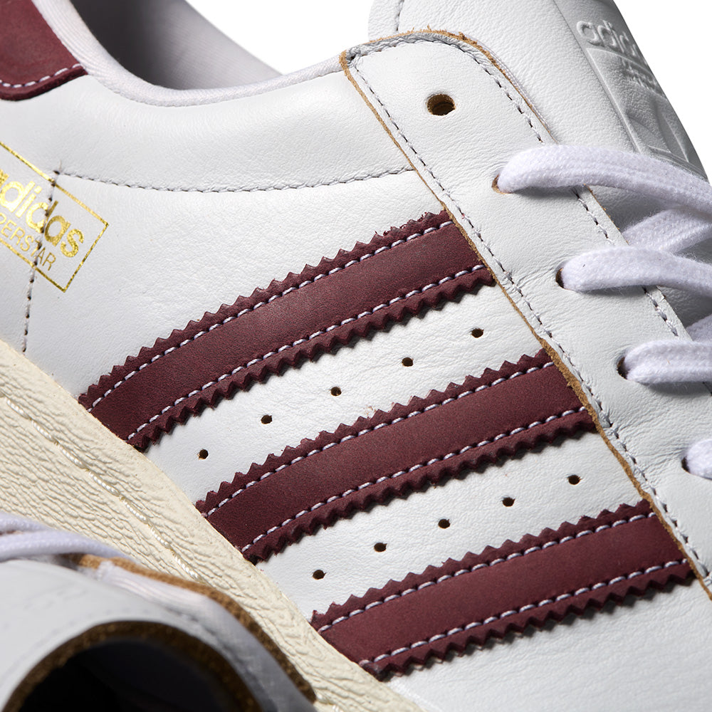 Close-up of the ADIDAS SUPERSTAR 80 ADV in Crystal White, featuring shadow red stripes, gold branding, perforations, and a textured off-white flexible cupsole.