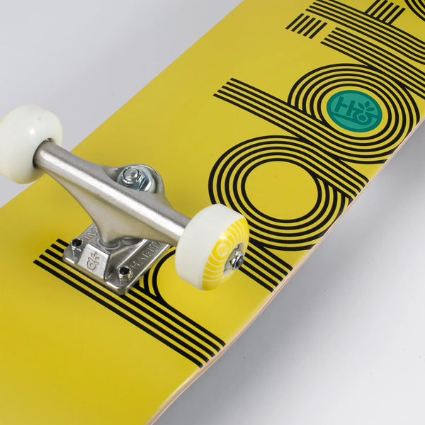 A close-up of a yellow skateboard with black graphic designs and a pair of silver trucks and white wheels, featuring the HABITAT ELLIPSE COMPLETE YELLOW design.