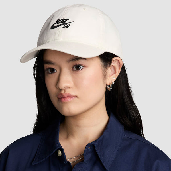 Woman wearing a white Nike SB Club Hat Sail and a navy blue shirt, looking to her left with a serious expression.