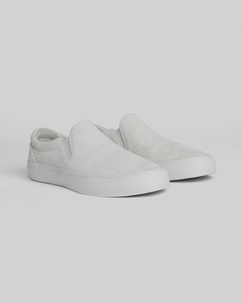 The WARSAW LTD STRANGER SLIP-ON | WHITE / WHITE features genuine pig suede in light gray, white rubber soles, and a minimal silhouette, displayed on a plain light background.