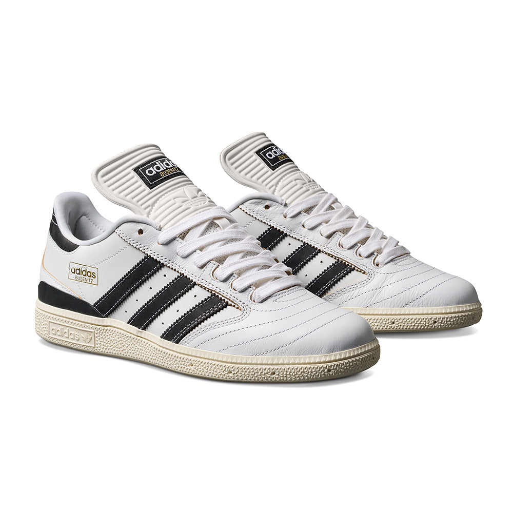 A pair of ADIDAS BUSENITZ sneakers in Crystal White, Black, and Off White featuring premium suede accents, beige soles, gold branding, and an Adiprene sockliner, displayed on a white background.
