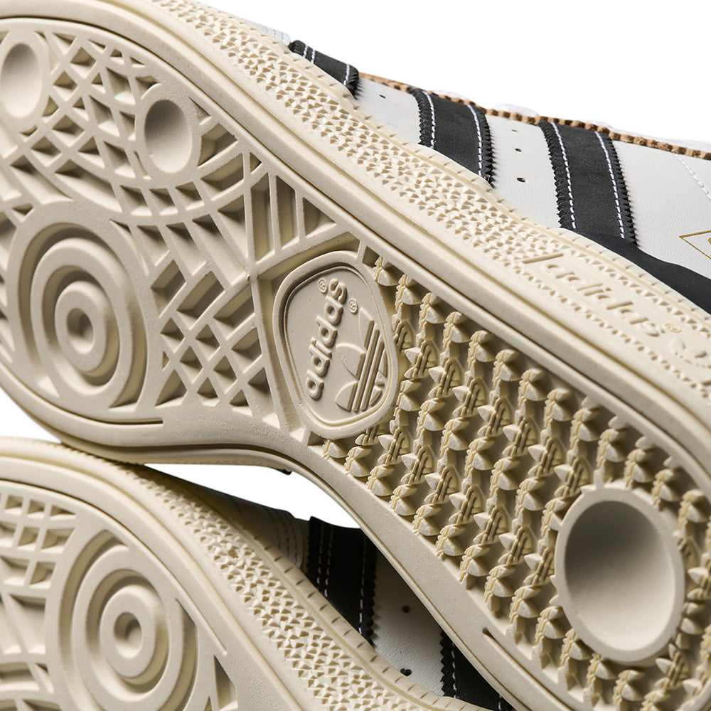 Close-up of the soles of ADIDAS BUSENITZ Crystal White/Black/Off White sneakers, highlighting the intricate tread patterns and iconic logo—designed for comfort.