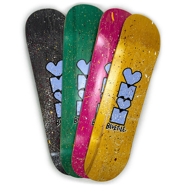 Four skateboard decks in black, green, pink, and yellow feature a blue graffiti-style design and the word "BUETTLE" on the bottom, showcasing Jay Croft Artwork for the exclusive Bluetile Skateboards HEART + SQUARE SPLATTER DECK.