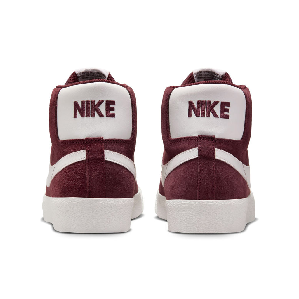 Back view of a pair of NIKE SB BLAZER MID BURGUNDY CRUSH / SUMMIT WHITE sneakers. The word "NIKE" is prominently displayed in maroon on the heel of each shoe.