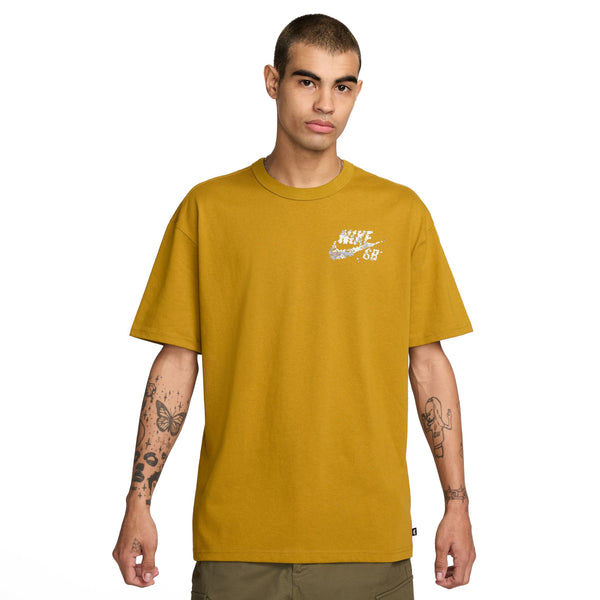 A young man wearing a mustard yellow Nike SB Yuto Tee Bronzine with a small logo on the chest stands against a white background.