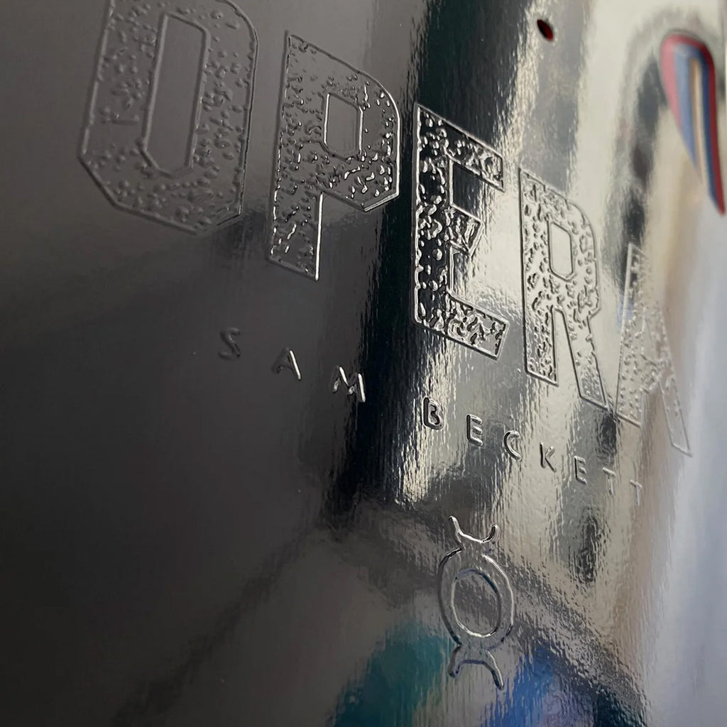 Close-up of the embossed text "OPERA BECKETT MELT" with a gender symbol on a wet metallic surface, reflecting light with hints of Popsicle Blue Foil.