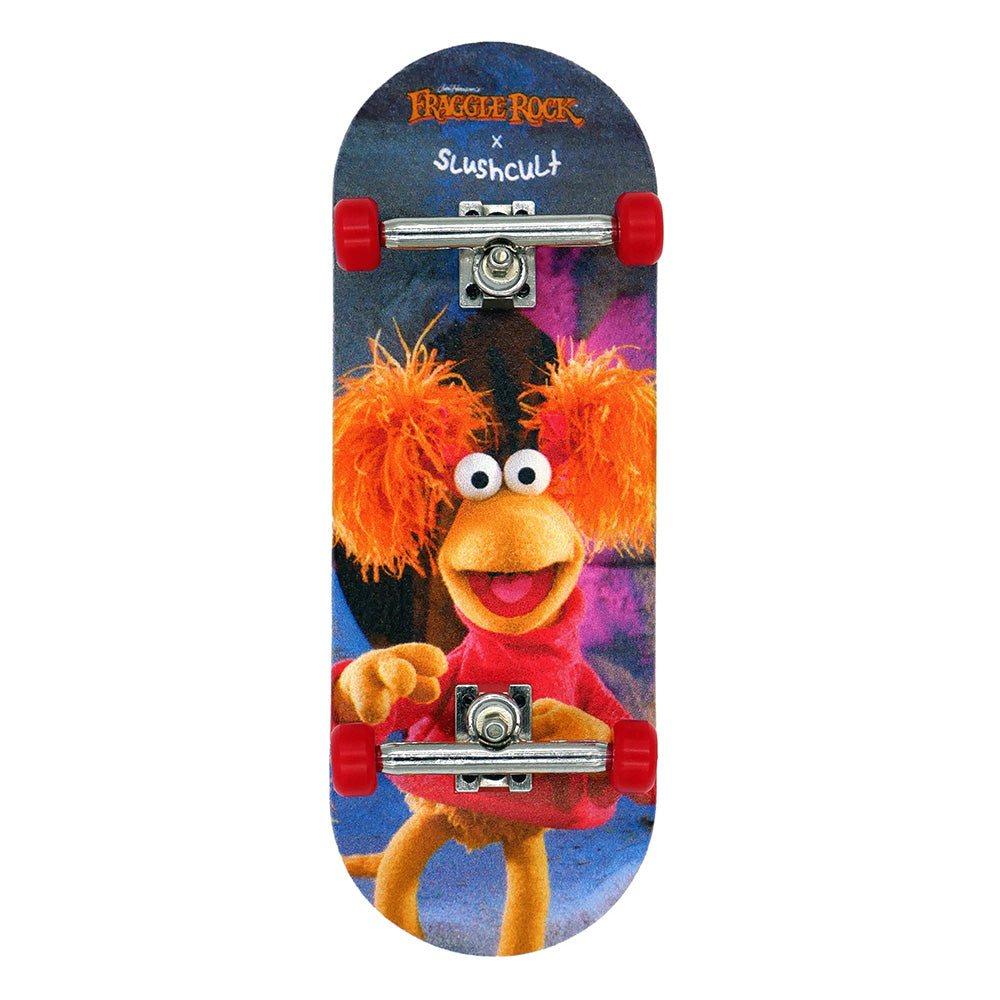 The SLUSHCULT X FRAGGLE FINGERBOARD RED features a Fraggle Rock character with orange hair and a red shirt, ideal for beginner fingerboarders, and comes with smooth polyurethane wheels.
