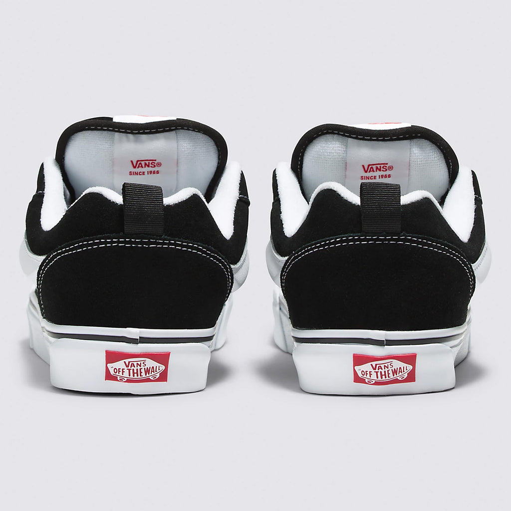 A pair of black and white VANS KNU SKOOL BLACK / TRUE WHITE sneakers viewed from the back. The red Vans "Off The Wall" logo is visible on the heel of each shoe. These shoes feature a Popcush insole for comfort and a vulcanized outsole for durability, making them perfect for both style and performance.