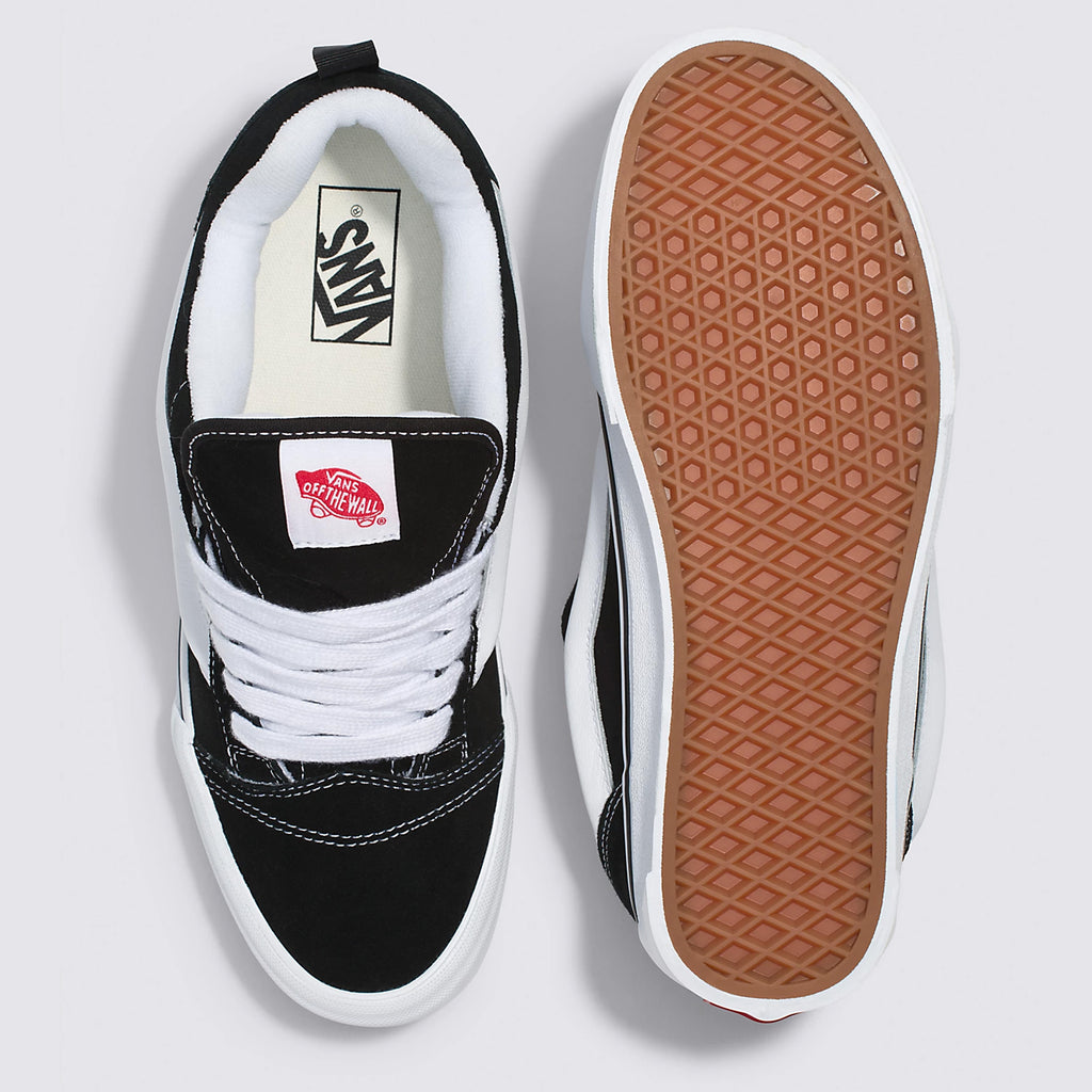A pair of black and white VANS KNU SKOOL BLACK / TRUE WHITE sneakers. The left shoe is shown top-down, displaying laces and the brand logo on the tongue, while the right shoe is shown from the sole, revealing the brown waffle pattern and vulcanized outsole.