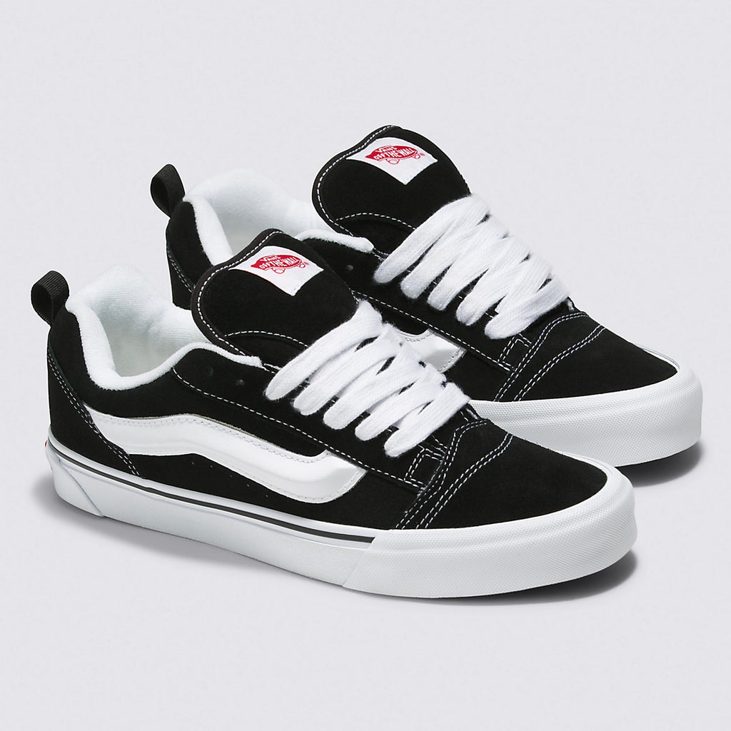 A pair of black and white low-top sneakers with a suede upper, white laces, and a wavy white stripe on the side, featuring a red logo on the tongue. Complete with Popcush insoles for added comfort, these VANS VANS KNU SKOOL BLACK / TRUE WHITE shoes are perfect for any skate enthusiast.
