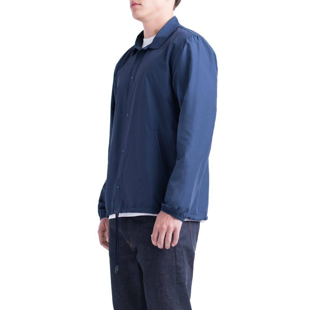 A person wears a plain navy blue HERSCHEL SUPPLY CO VOYAGE COACH PEACOAT over a white shirt and dark jeans, standing against a white background.