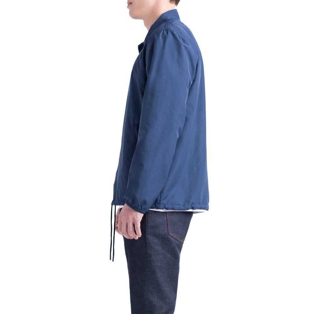 A person in profile wears a blue HERSCHEL SUPPLY CO VOYAGE COACH PEACOAT by HERSCHEL, made from water-resistant ripstop, styled with a white shirt and dark jeans against a white background.