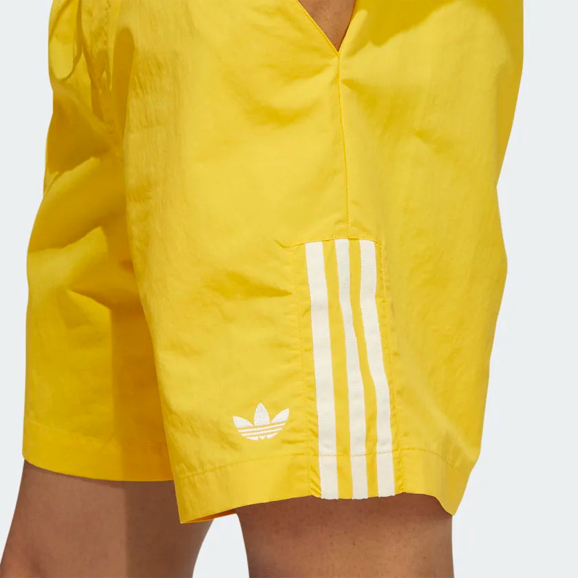 A person is wearing ADIDAS WATER SHORT BOLD GOLD/CREAM WHITE—bright yellow, gender-neutral skateboarding water shorts with white stripes and an ADIDAS logo on the left side.