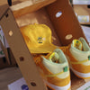 A pair of yellow BLUETILE SMOKE SQUARES NYLON 6 PANEL YELLOW sneakers and a hat in a cardboard box. The yellow hat is made of nylon and has a six-panel design, perfect for any casual or sporty outfit. Alongside Bluetile Skateboards.
