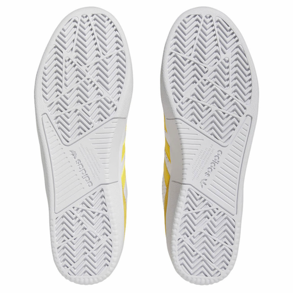 A bottom view of the ADIDAS TYSHAWN LOW shoes in bold gold, white, and black reveals textured rubber soles with herringbone tread patterns for grip and stability. The soles also feature prominent branding logos.