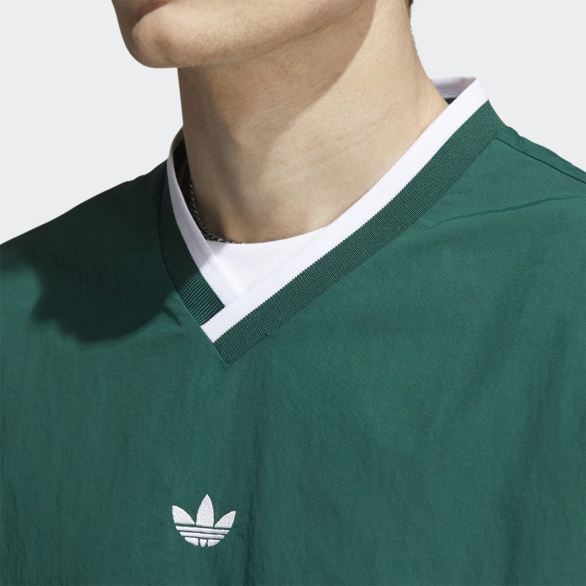 A person wears the ADIDAS WIND PULLOVER COLLEGIATE GREEN / WHITE, featuring a white logo and V-neck collar. The image spotlights the upper chest and neck, emphasizing its sporty, vintage golf windbreaker style.