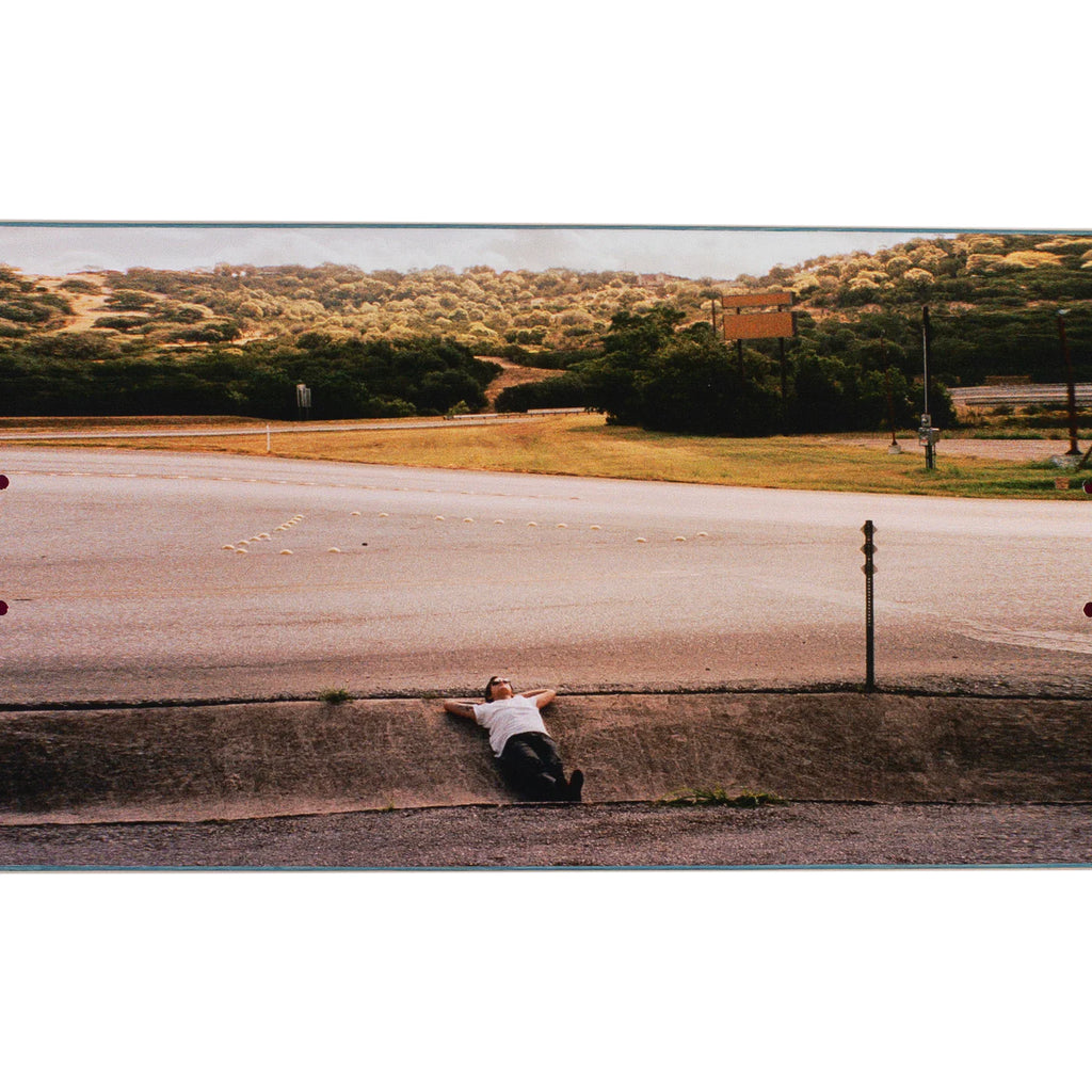 A man is laying on the side of a road, surrounded by various stains, holding the FUCKING AWESOME DYLAN PHOTO BY DILL.