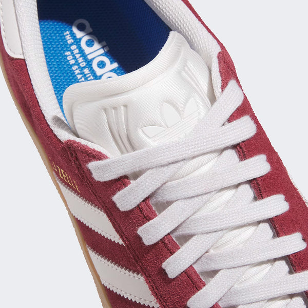 Close-up of the ADIDAS GAZELLE ADV in Shadow Red, Crystal White, and Gold Metallic—featuring signature white stripes, white laces, and a blue Adidas-branded insole; a classic from ADIDAS known for skate-ready durability.