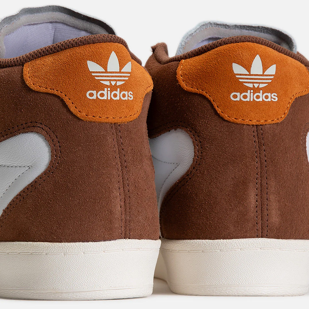 Close-up of the back of two ADIDAS SUPERSKATE ADV sneakers in Preloved Brown and Rustic Orange with white soles, displaying the Adidas trefoil logo and text on the heel tabs.