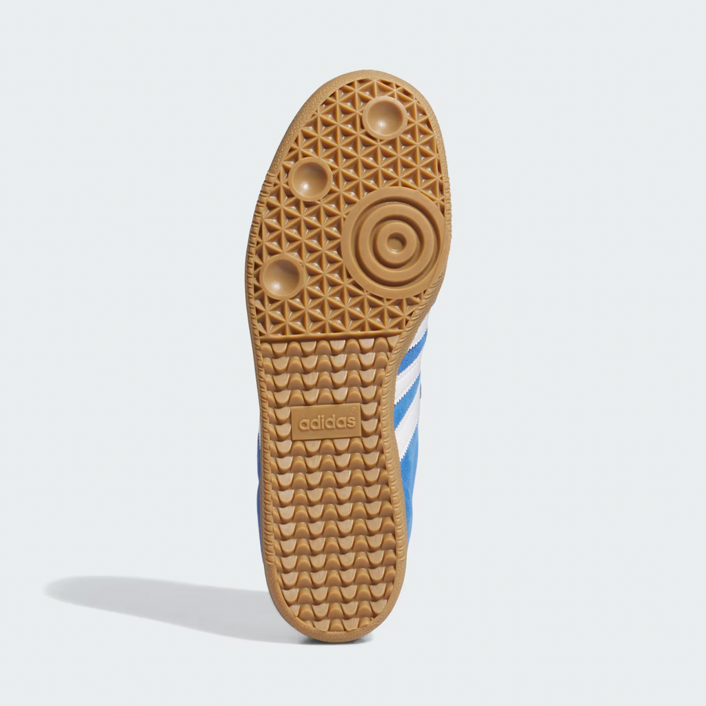The image features the cupsole of the Adidas Samba ADV in Blue Bird, Cloud White, and Gold Metallic, with a textured rubber pattern and brand logo centrally placed. It also boasts Adituff toe reinforcement for enhanced durability.