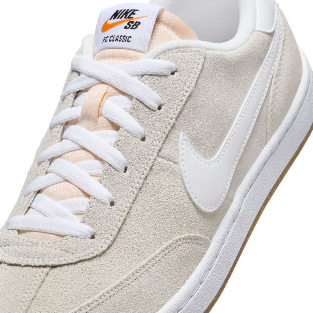 A close-up of the NIKE SB FC CLASSIC SUMMIT WHITE / SUMMIT WHITE sneaker showcases its cargo khaki design with white laces and an iconic white swoosh logo. It features a sleek white sole and an orange logo on the tongue, making it perfectly designed for skateboarding enthusiasts.