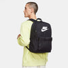 A person in a light yellow sweatshirt carries a black Nike Heritage Backpack, known for its durable, water-resistant material and convenient compartments.