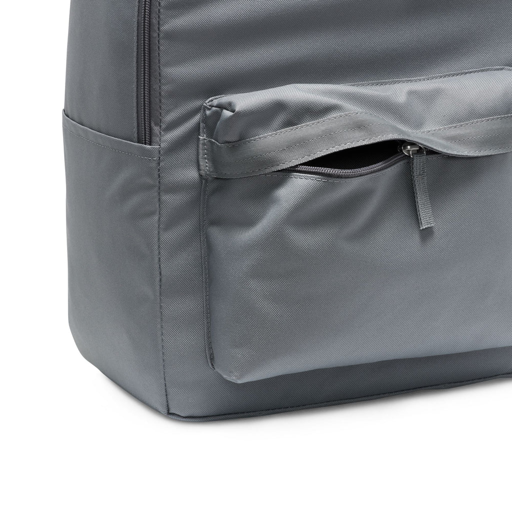 The NIKE HERITAGE BACKPACK in Smoke Grey/White is made from recycled polyester and includes a front zippered pocket, a side pocket, and a laptop sleeve.