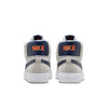 Rear view of a pair of NIKE SB BLAZER MID WOLF GREY / MIDNIGHT NAVY sneakers in light grey and blue with "NIKE" in orange on the back, featuring Zoom Air cushioning.