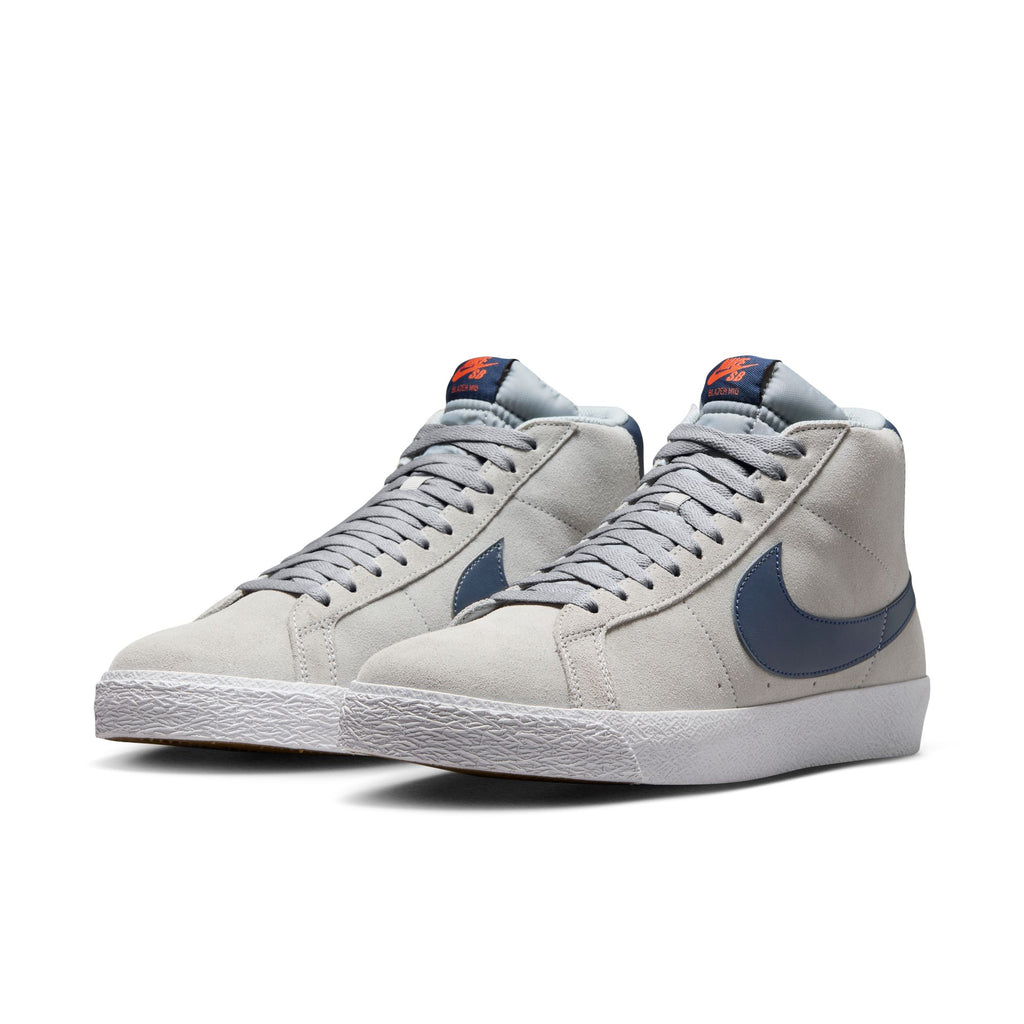 A pair of NIKE SB BLAZER MID WOLF GREY / MIDNIGHT NAVY high-top sneakers features a light gray color with blue swooshes, white soles, gray laces, orange branding on the tongue, and Zoom Air cushioning designed for skateboarders.