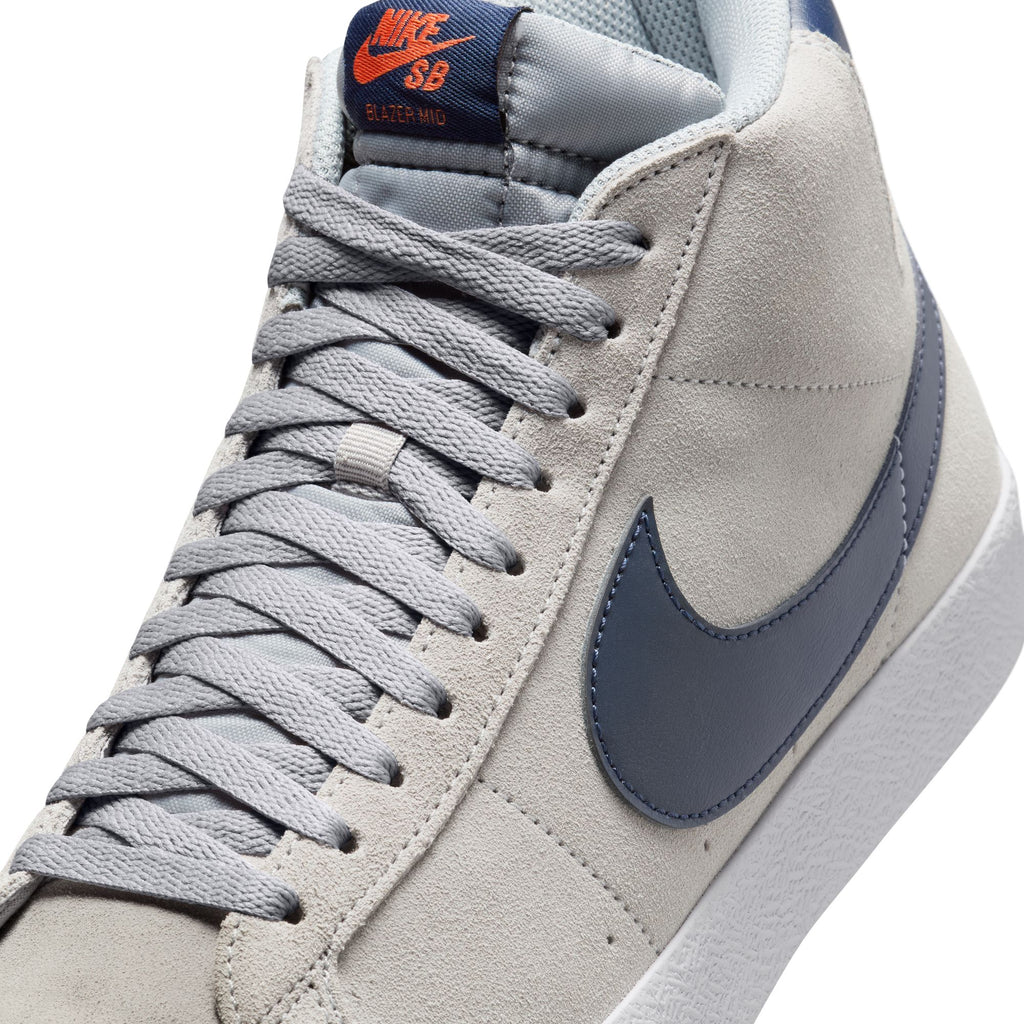 Close-up view of the NIKE SB BLAZER MID WOLF GREY / MIDNIGHT NAVY high-top sneaker featuring light gray color, gray laces, a blue swoosh logo on the side, and orange "Nike SB Blazer Mid" branding on the tongue, with Zoom Air cushioning ideal for skateboarders.