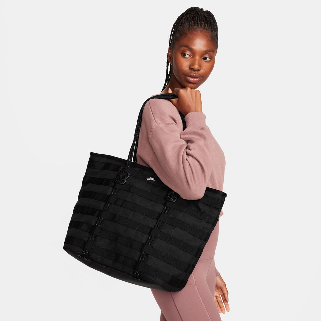 A woman in a mauve sweatshirt and leggings carries the Nike RPM Tote (26L) Black over her shoulder, looking to the side against a plain background.