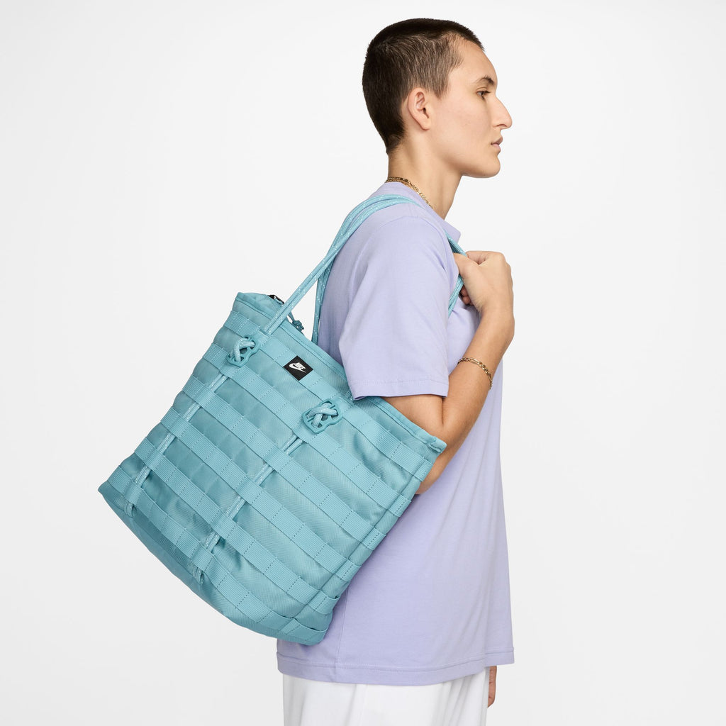 Person in a lavender t-shirt carries the NIKE RPM TOTE (26L) in Denim Turquoise / Black / Summit White by Nike on their shoulder, standing against a plain white background.