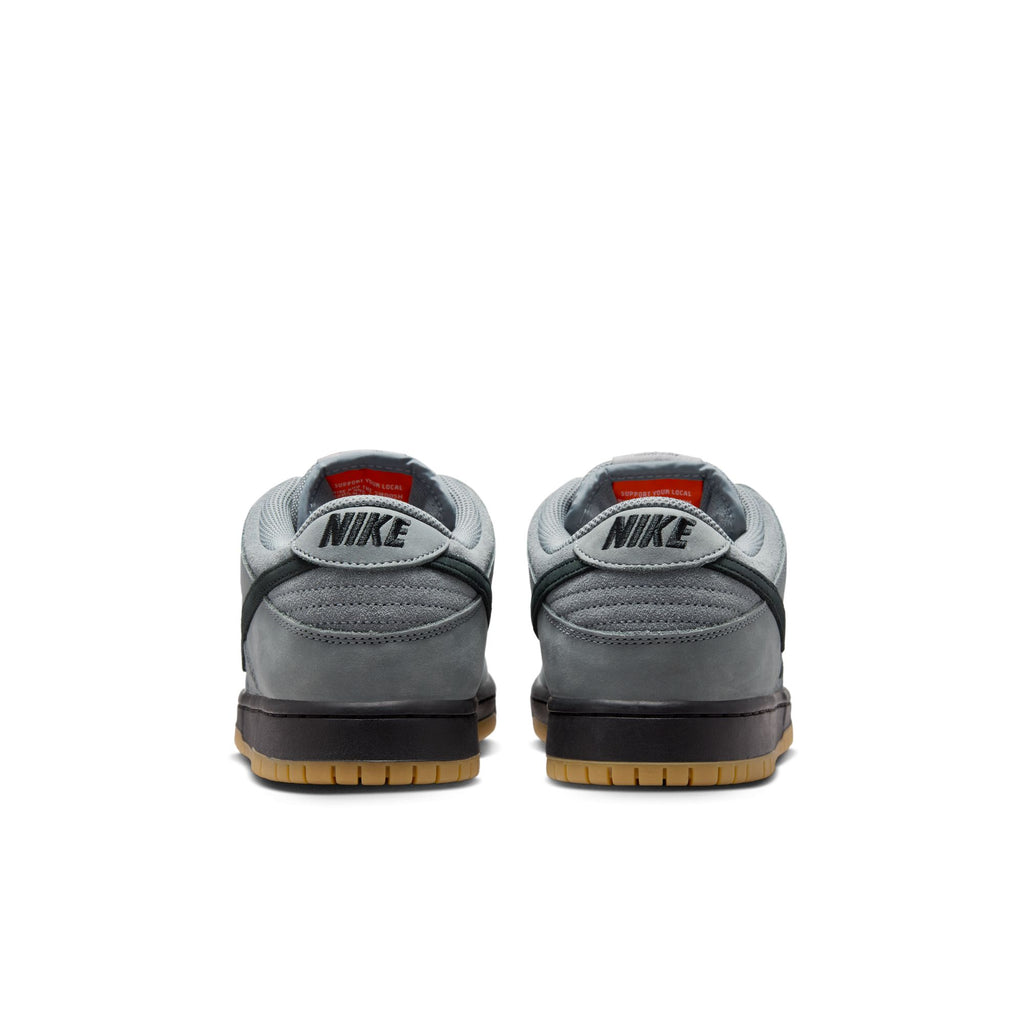 A pair of NIKE SB DUNK LOW PRO in cool grey and black, viewed from the back, feature black “NIKE” text on the heel and light brown rubber soles, echoing the classic skateboarding style by Nike.