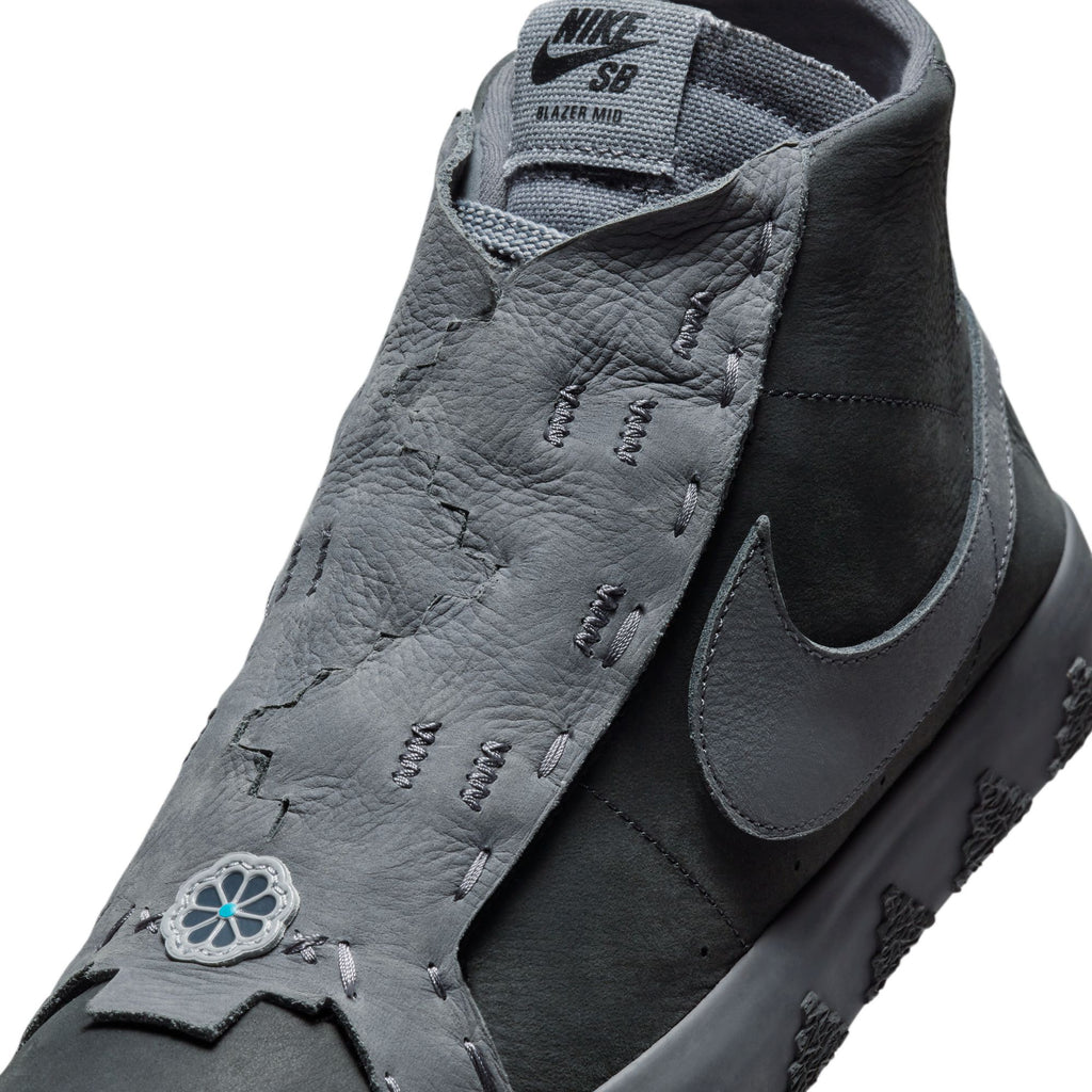 Close-up view of a gray and black nike NIKE SB X DI'ORR GREENWOOD BLAZER MID DECON ANTHRACITE / DARK GREY SMOKE skate shoe with a unique textured upper and a floral emblem near the toe. The sneaker features visible stitching, a prominent Nike swoosh logo, and Zoom Air cushioning for added comfort.