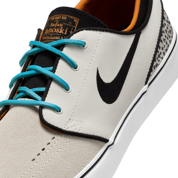 Close-up image of the NIKE SB ZOOM JANOSKI OG+ skateboard shoe in Electric Phantom/Chlorine Blue-Black, featuring a white upper with a black Nike swoosh, blue laces, orange inner lining, and speckled heel detail. The tongue label reads "Stefan Janoski." Designed for performance, this Nike SB shoe includes Zoom Air cushioning for unparalleled comfort while skateboarding.