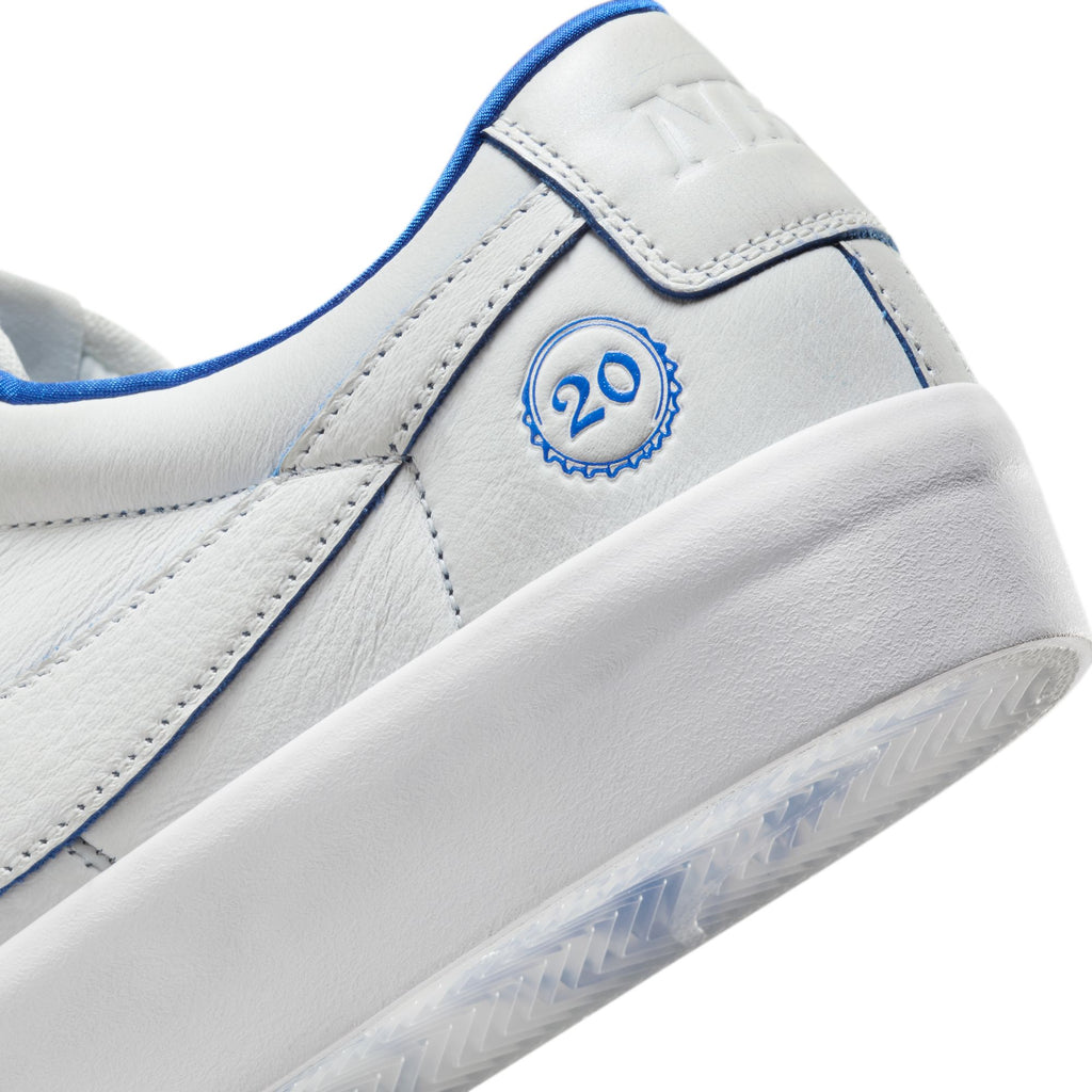 The NIKE SB BLAZER LOW PRO GT "20 YEARS" in Summit White/Game Royal showcases a close-up of a white sneaker with blue trim, adorned with the "20" logo on the side. This design, inspired by Grant Taylor, features Air Zoom cushioning for exceptional style and comfort.