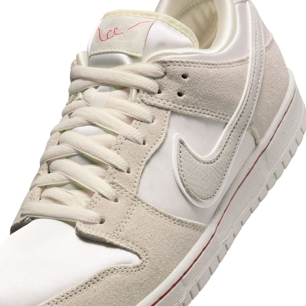 Close-up of a white sneaker with beige suede accents, featuring a prominent Nike Swoosh logo on the side and red stitching detail on the sole, inspired by the NIKE SB 'CITY OF LOVE' DUNK LOW PRO WHITE / RED from nike.