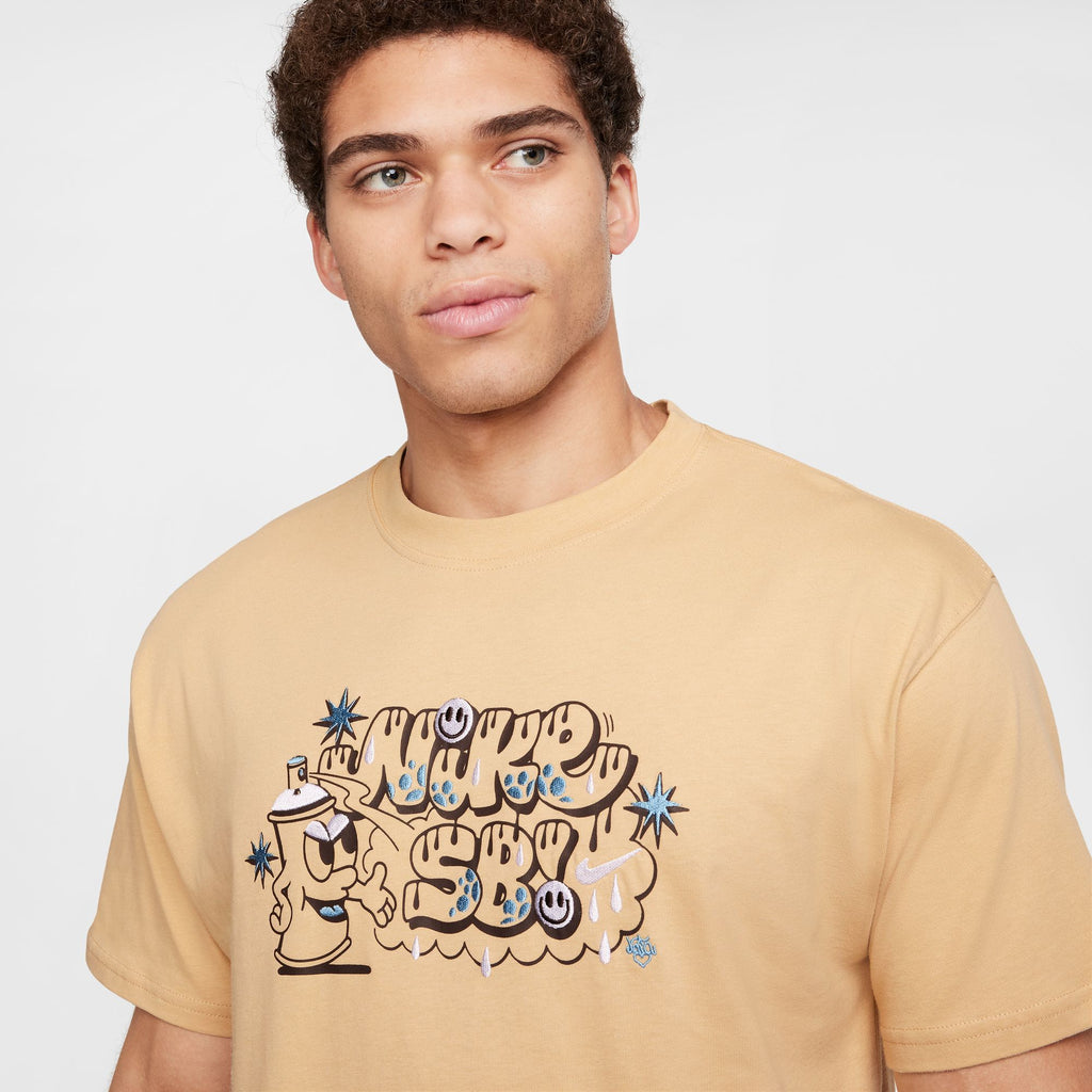 Someone wearing the NIKE SB Max90 Spray Can Skate Tee in Sesame, a beige cotton t-shirt featuring vibrant graffiti-inspired text and designs with a glow-in-the-dark effect.