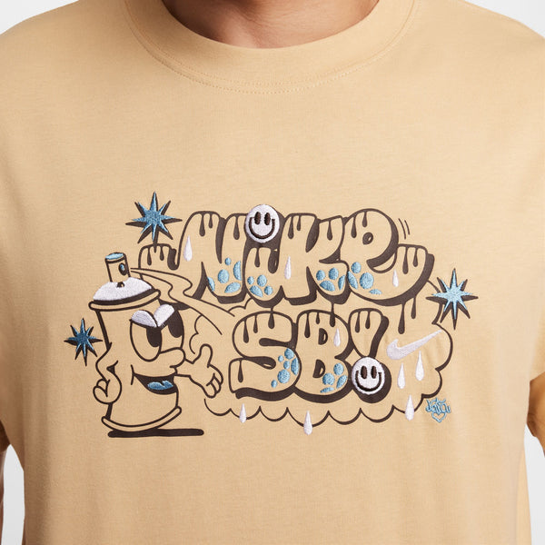 A person wears the "NIKE SB MAX90 SPRAY CAN SKATE TEE SESAME" by Nike, a beige T-shirt adorned with a spray paint can character and graffiti-like "NIKE SB" text. Glow-in-the-dark stars and smiley faces enhance this striking design.