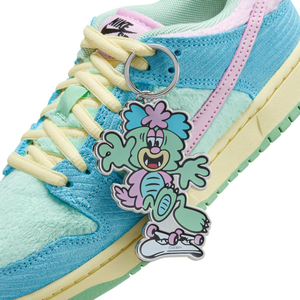A close-up of the NIKE SB DUNK LOW PRO VERDY PS in Blue Gaze, Arctic Pink, and Bicycle Yellow showcases its light blue and pastel hues. The sneaker features a playful cartoon keychain attached to the laces, making it perfect for little kids.