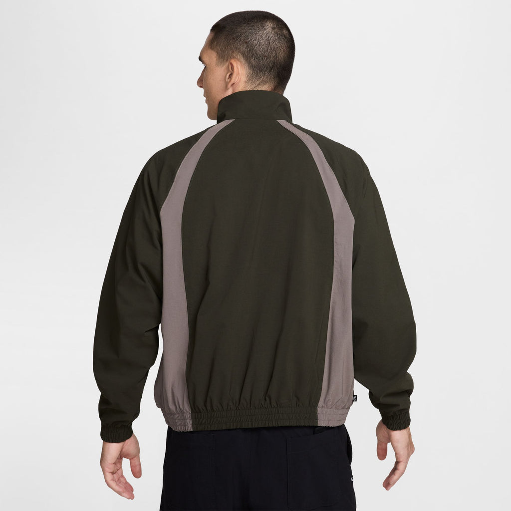 A person is shown from behind wearing a NIKE SB Full-Zip Woven Skate Jacket in Sequoia and Cave Stone, standing against a plain background.