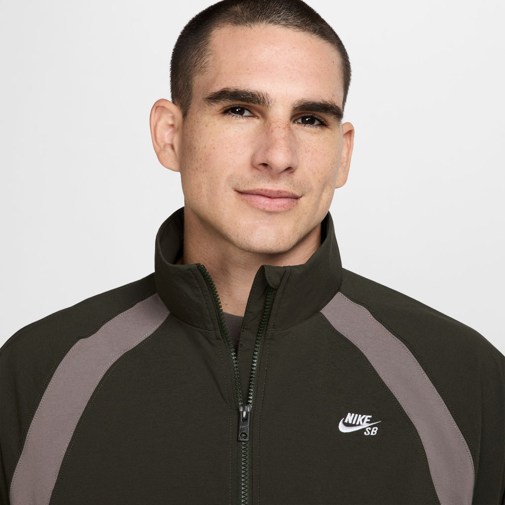 A person with short hair is wearing a dark high-collar Nike SB Full-Zip Woven Skate Jacket in Sequoia/Cave Stone/White that resembles a stylish skate hoodie.